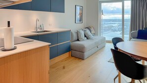 Une cuisine moderne avec un évier, un coin repas comprenant une table et des chaises, un canapé gris agrémenté de coussins et une grande fenêtre offrant une vue sur la ville.