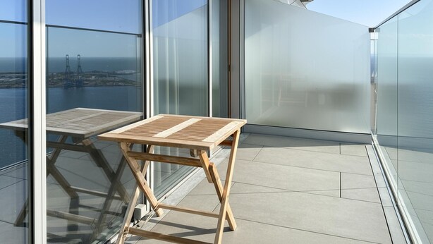 Un balcon avec une table et des chaises en bois, de grandes fenêtres en verre et une vue sur la mer et le paysage urbain.