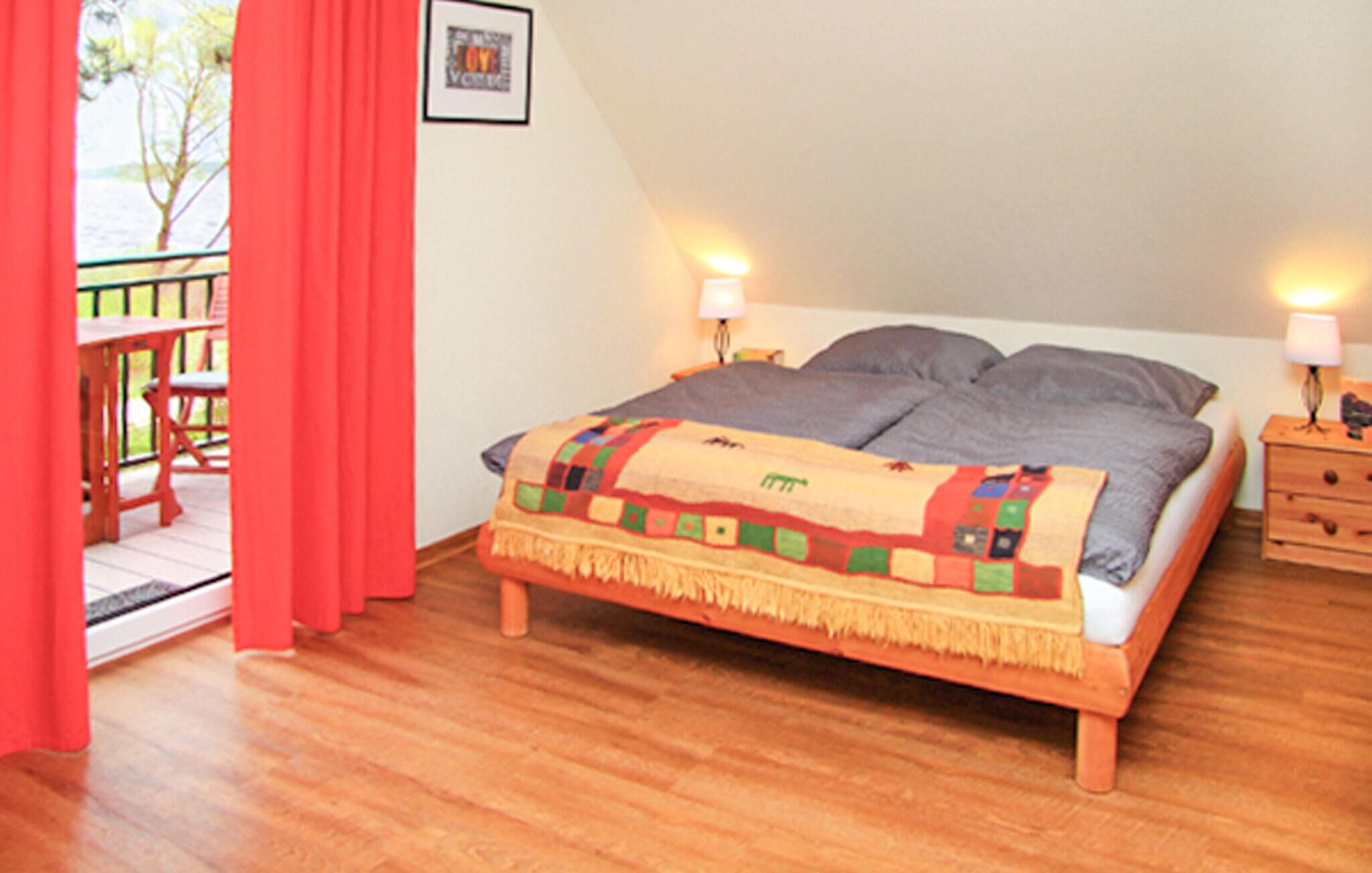 A bedroom with a wooden bed, two bedside lamps, a red curtain, and a small table with chairs outside.