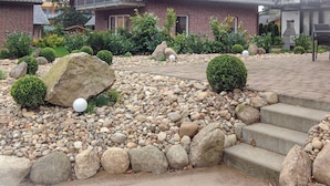 Un jardín ajardinado con diversas rocas y arbustos, un sendero pavimentado y un conjunto de escalones de concreto que llevan a un edificio.