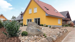 Una casa amarilla con techo rojo, una roca grande con la inscripción 'Haus am See' y un camino de grava.