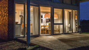 Een modern huis met grote glazen deuren die leiden naar een eetgedeelte, een zichtbare keuken en een terras met tuinmeubilair.