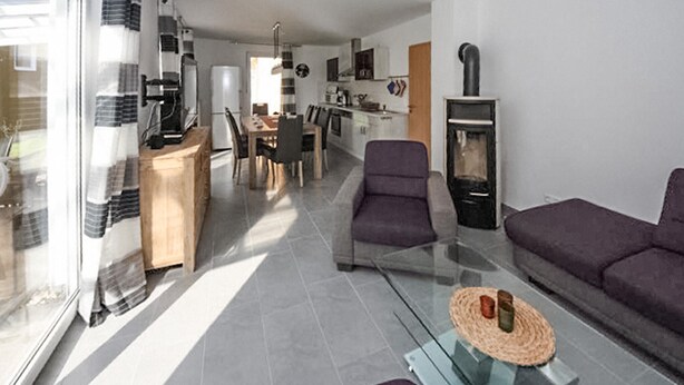 En moderne stue med et glasbord, en sofa og et spiseområde, hvor et køkken ses i baggrunden.