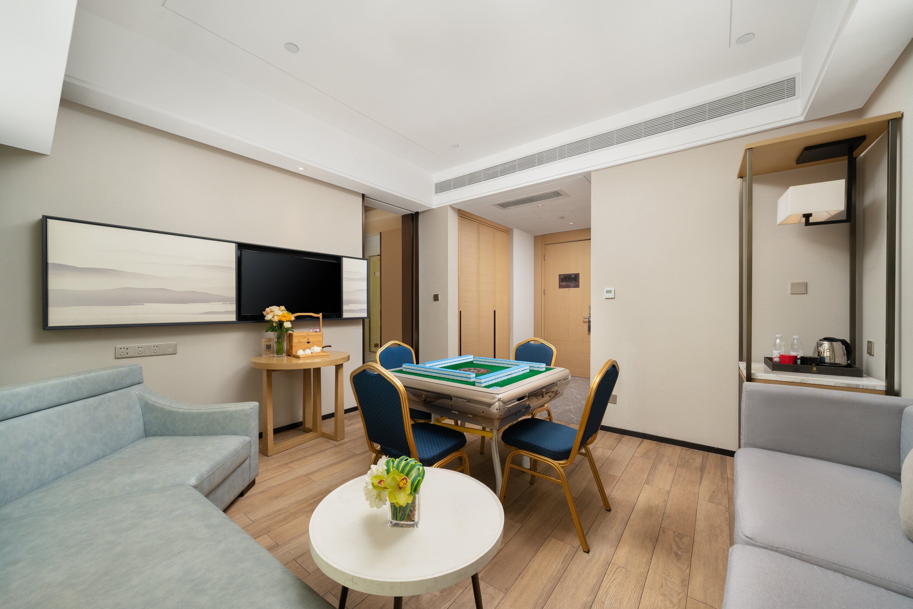 A modern hotel room with a sofa, a dining table with chairs, a flat-screen TV mounted on the wall, and a small side table with a vase of flowers.
