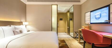 A hotel room with a large bed, a desk with a chair, a television, and a glass door leading to another room.