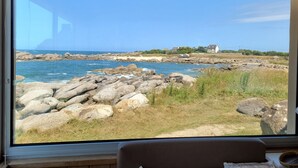 Blick aus einem Zimmer mit großem Fenster auf eine felsige Küste und Häuser in der Ferne.