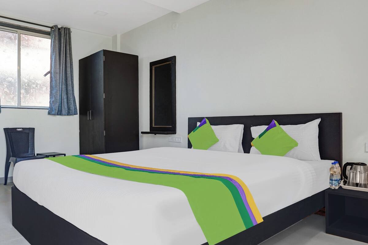 A hotel room with a large bed, a chair, a wardrobe, a nightstand, and a window with curtains.