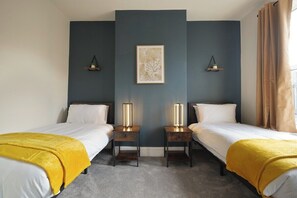 Ein Hotelzimmer mit zwei Betten, einer dunkelblauen Akzentwand, einem gerahmten Kunstwerk, Nachttischlampen und einem Fenster mit Vorhängen.
