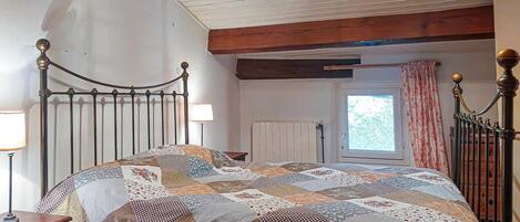 A bedroom with a metal bed frame, a quilted bedspread, a nightstand with a lamp, a radiator, and a window with curtains.