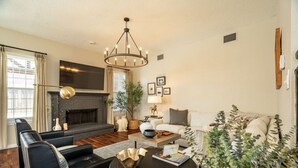 A living room with a fireplace, black leather sofas, a round coffee table, and a chandelier. - Designer-Styled Modern Farmhouse w/ Heated Pool (Allen)