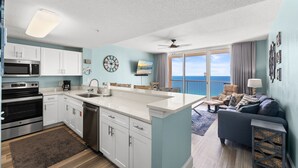 A modern kitchen with white cabinets, a built-in microwave, and a dishwasher. There is a large island with a sink and a ceiling fan. The living area features a gray sofa, a coffee table, and a view of the ocean through large windows. - Majestic 1710 | Beach-Front Gulf Views - 2BR/2BA (Panama City Beach)