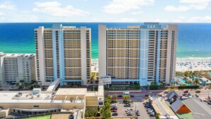 A beachfront hotel with multiple high-rise buildings, a parking area, and a beach with umbrellas and palm trees. - Majestic 1710 | Beach-Front Gulf Views - 2BR/2BA (Panama City Beach)