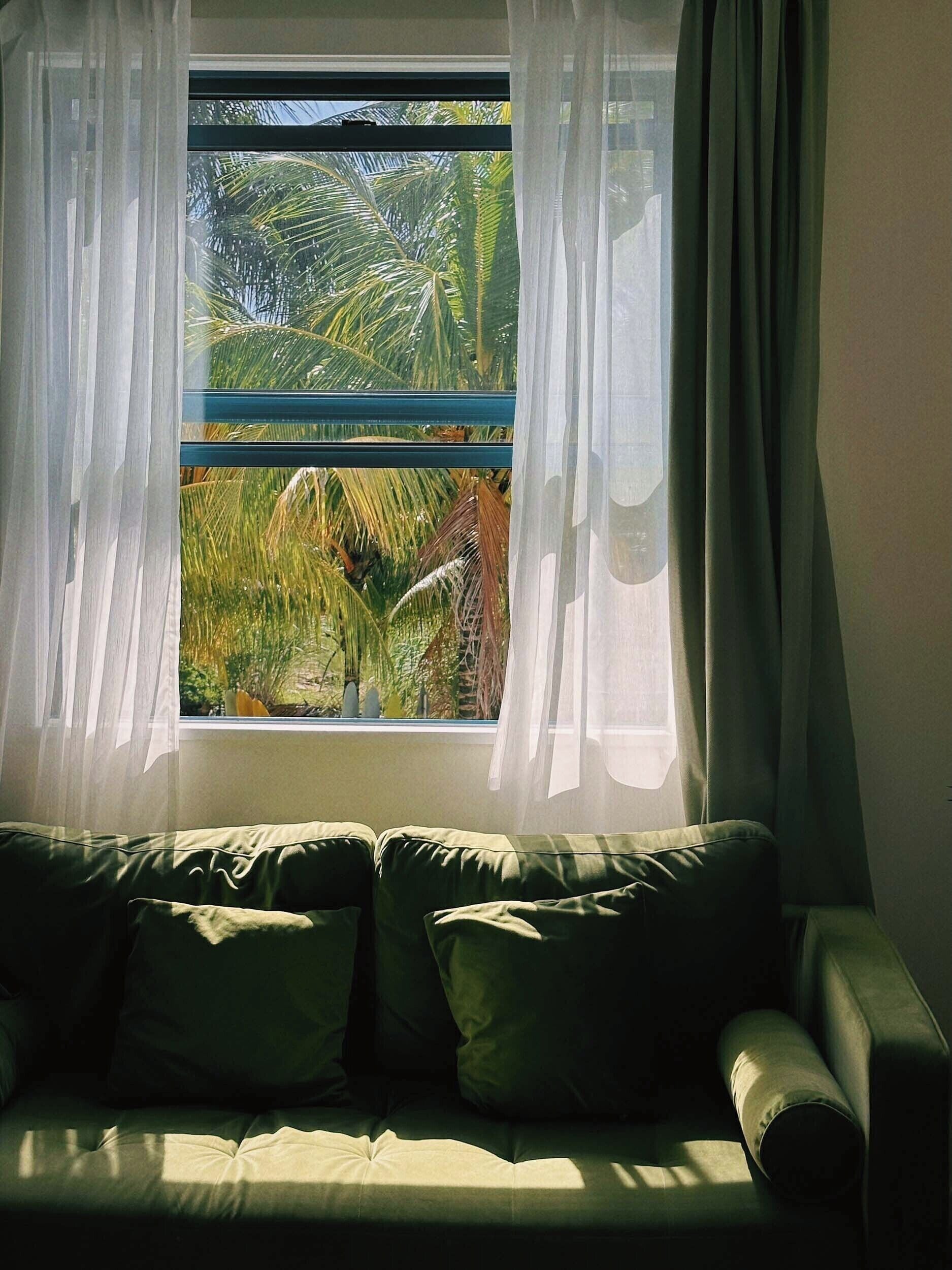 Een kamer met een raam, een bank en uitzicht op palmbomen.