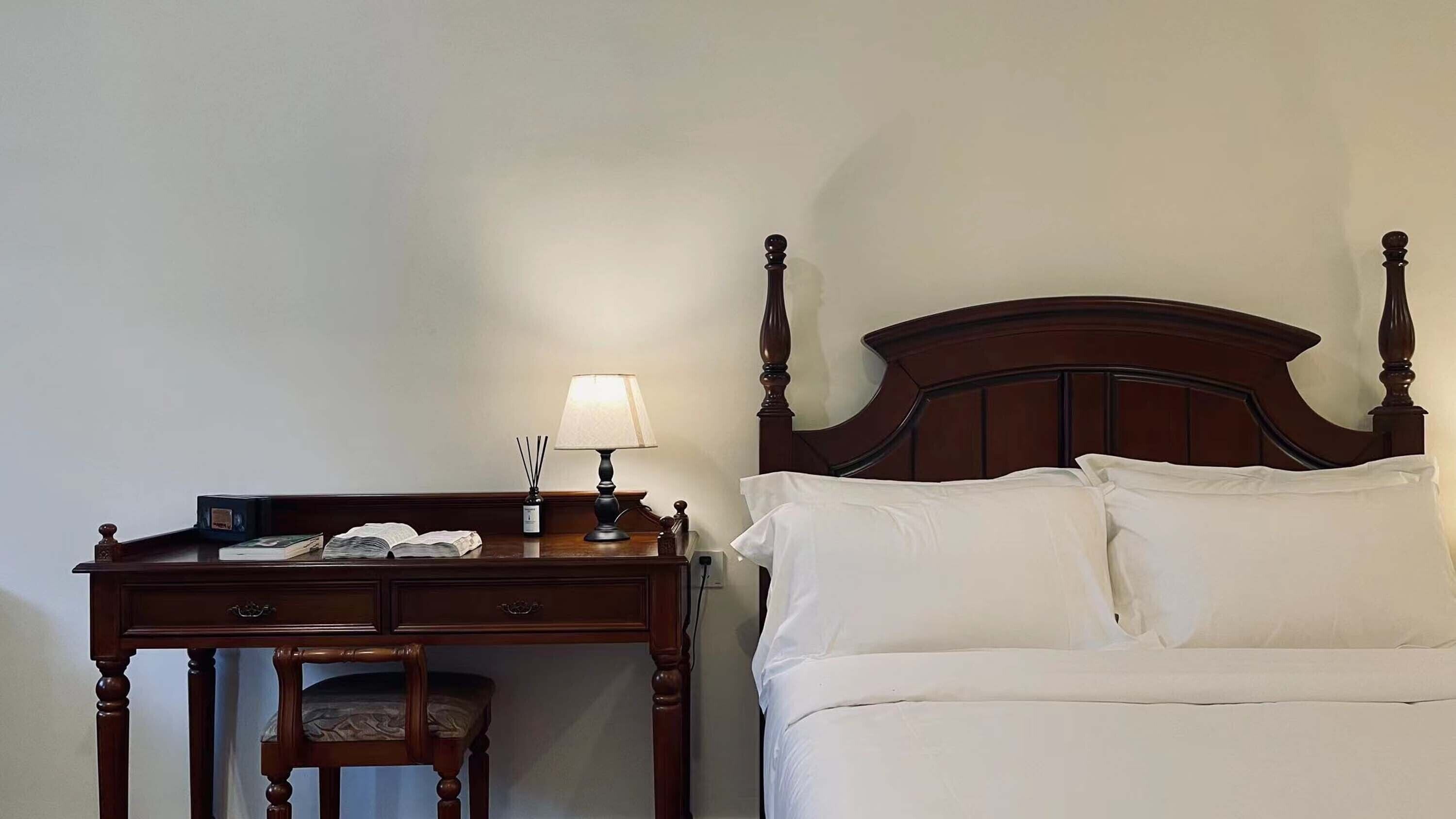 Una habitación de hotel con una cama de madera, almohadas blancas, una mesita de noche de madera con una lámpara y una silla.