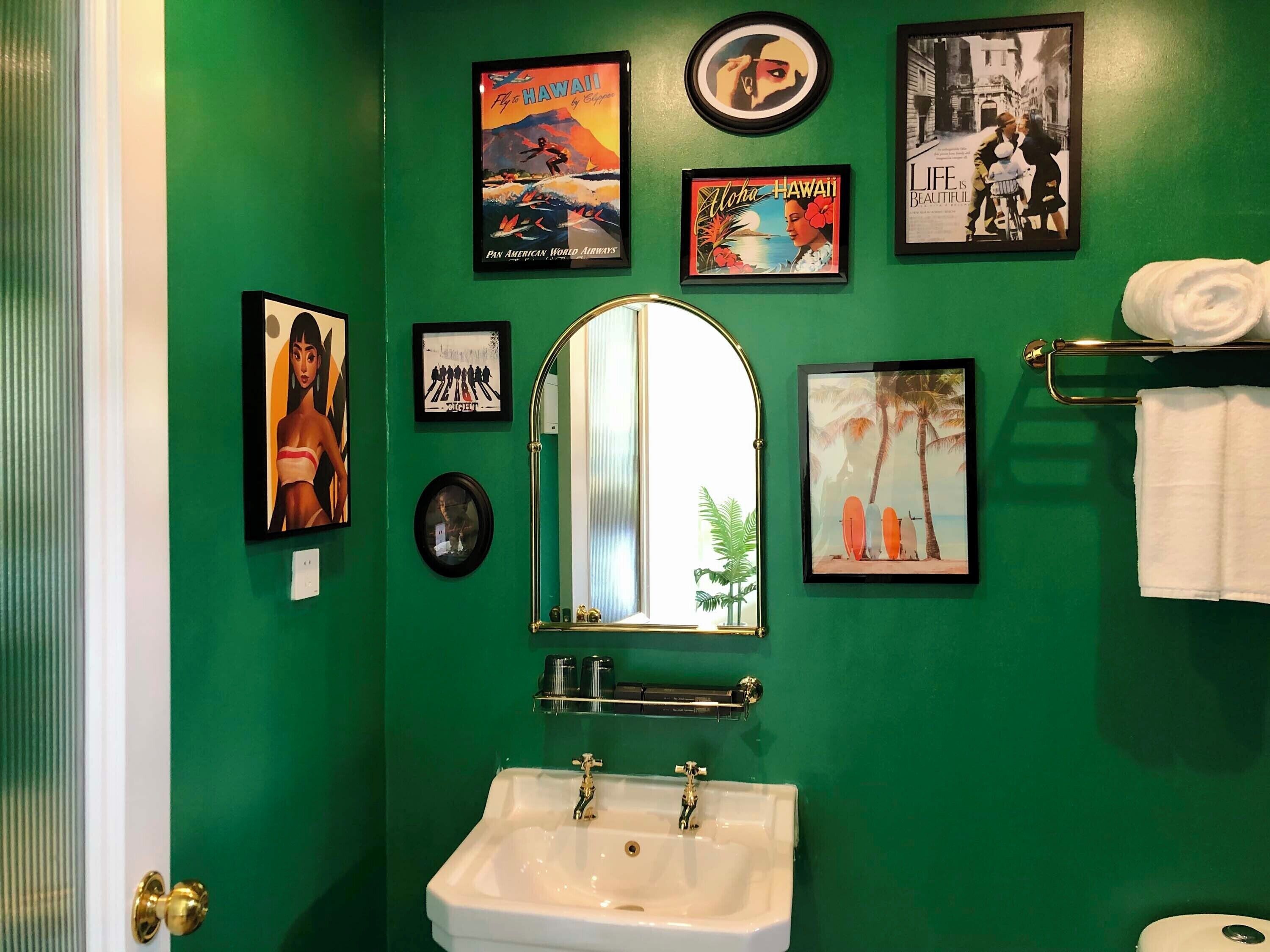 Un baño con pared verde, un lavamanos blanco y varias fotografías enmarcadas, incluyendo carteles de viaje y un espejo.