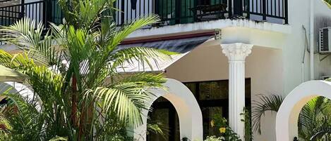 Una casa de dues plantes amb un balcó, finestres grans i un jardí ple de plantes tropicals.