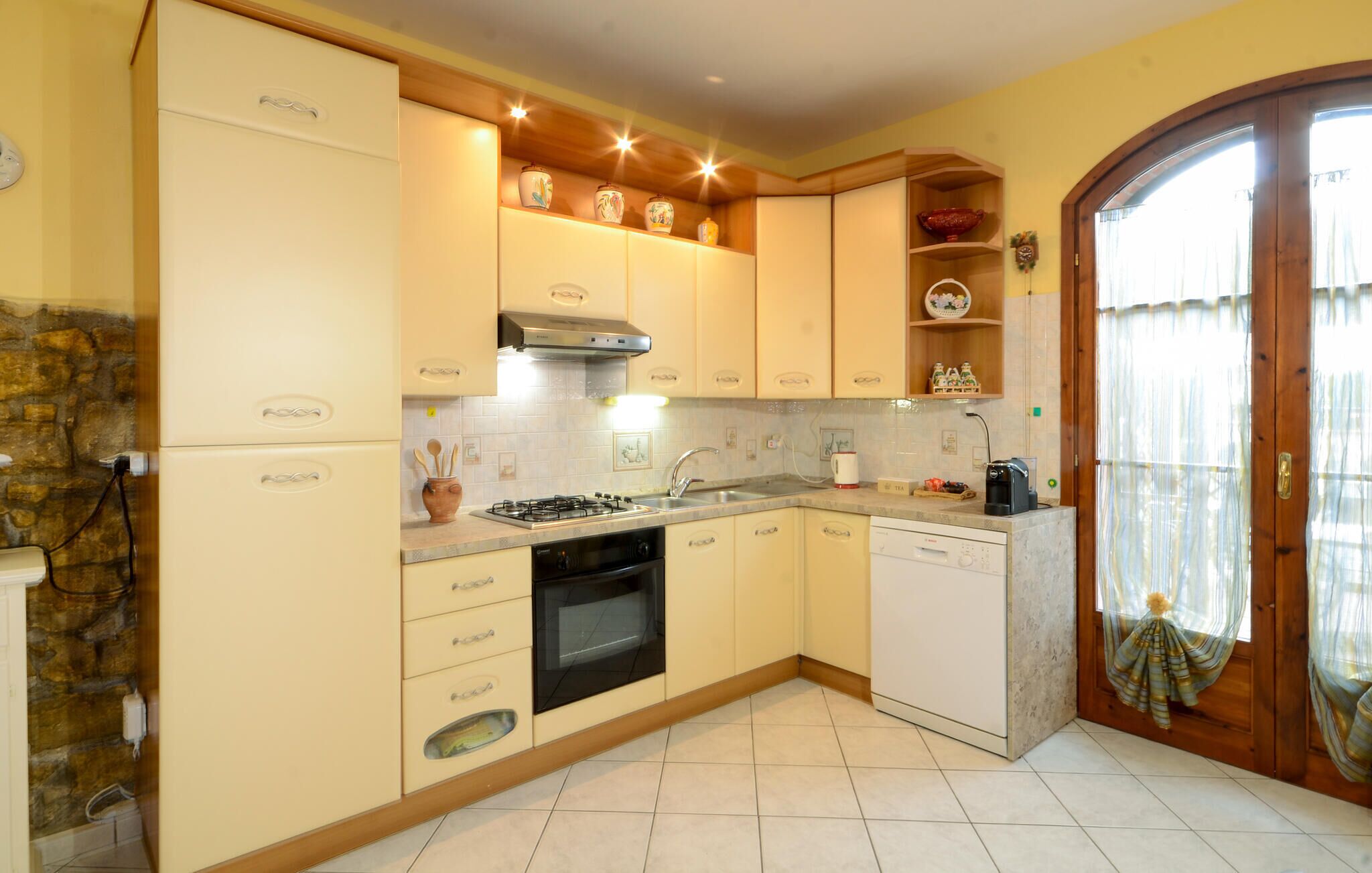 A compact kitchen with a gas stove, oven, and built-in dishwasher. It features light-colored cabinets and a stone wall on one side.