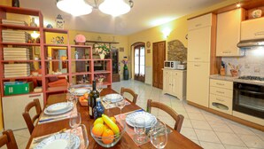 Un comedor con una mesa de madera dispuesta para cuatro personas, rodeada de sillas. Hay estanterías con libros y objetos decorativos, una cocina con electrodomésticos y un comedor con una araña.