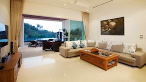 A modern living room with a sofa, coffee table, and a TV. There is a large window with a view of greenery and buildings. - Kamala Falls 3BR Seaview w Private Pool (Kamala)