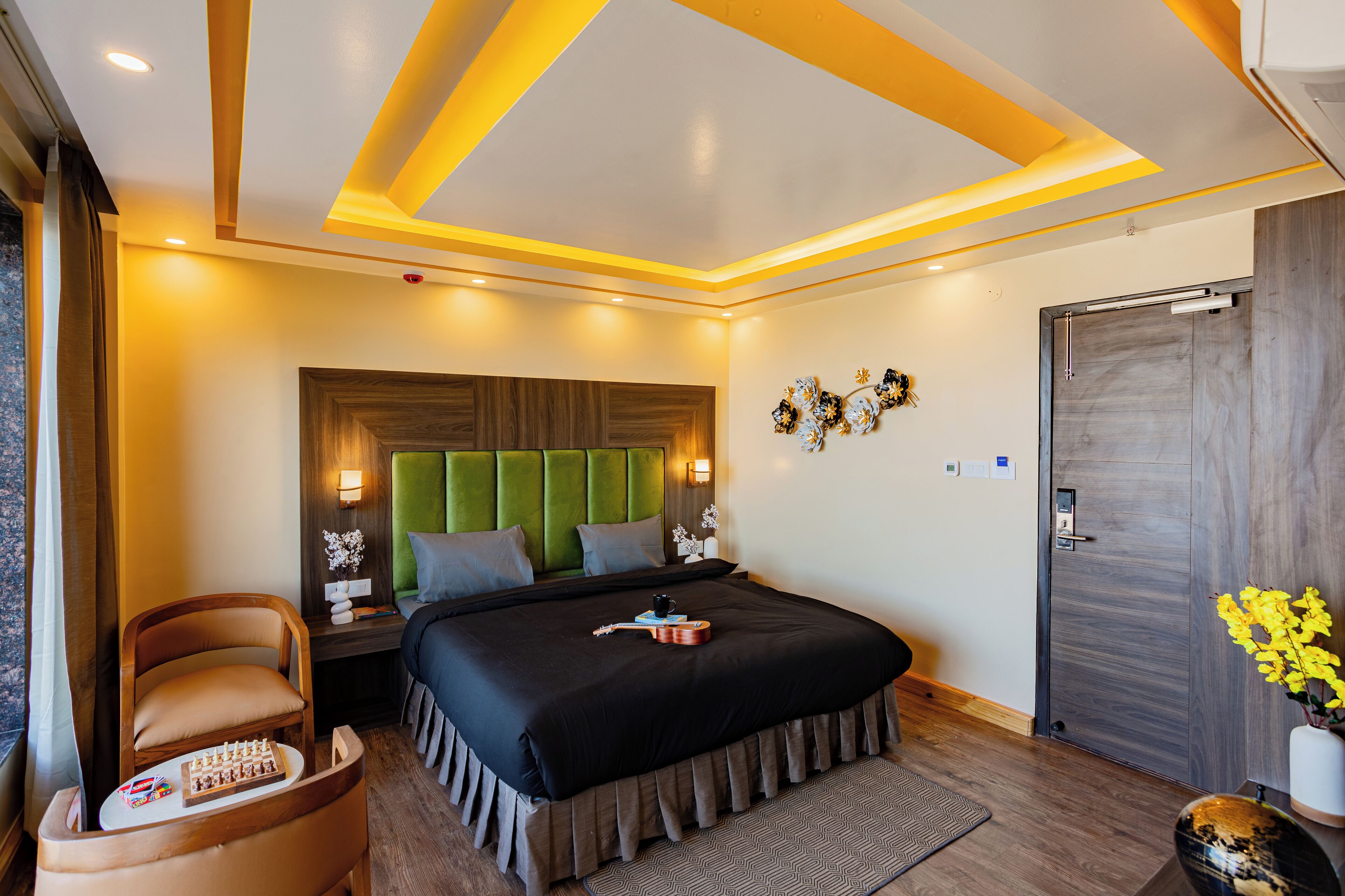 A modern hotel room with a large bed, a green upholstered headboard, a wooden nightstand, and a chair with a chessboard.