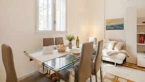 Ein moderner Essbereich mit einem Glastisch, einer weißen Tischdecke und Blick ins Freie. Ein Sofa und ein Bett befinden sich im angrenzenden Raum.