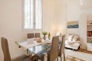 A modern dining area with a glass table, white tablecloth, and a view of the outdoors. There is a sofa and a bed in the adjacent room. - Lemon Tree House – Simple, Bright, and Peaceful (Bari)