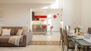 Ein modernes Wohnzimmer mit einem braunen Sofa, einem Esstisch für vier Personen und einer Küche im Hintergrund mit roten Akzenten.