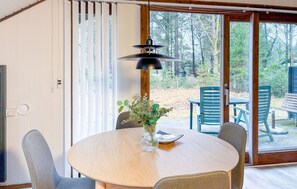 Una mesa redonda de madera con un florero de cristal transparente, rodeada por cuatro sillas, en una habitación con un amplio ventanal corredero que da a un espacio exterior con sillas y una mesa.