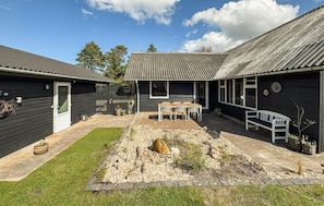 Il s’agit d’une propriété résidentielle dotée d’un patio, d’une table et de chaises en bois, d’un banc blanc et d’un jardin agrémenté de rochers et de plantes.