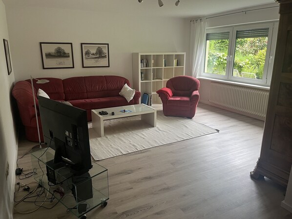 Ein Wohnzimmer mit einem roten Sofa, einem weißen Couchtisch, einem roten Sessel, einem Fernseher auf einem Glastisch und einem Bücherregal.