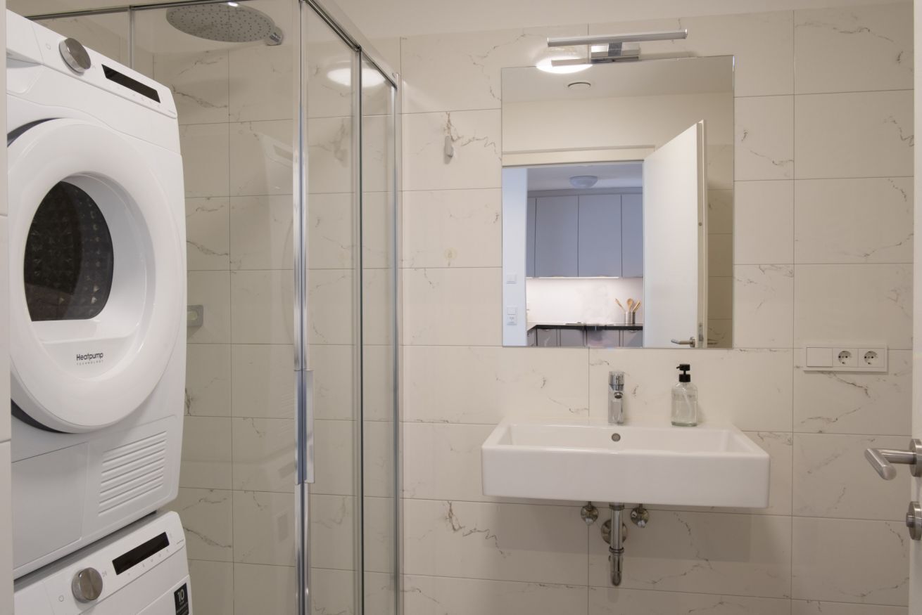 Ein modernes Badezimmer mit eingebauter Waschmaschine und Trockner, einer Glasdusche und einem Waschbecken mit Spiegel darüber.