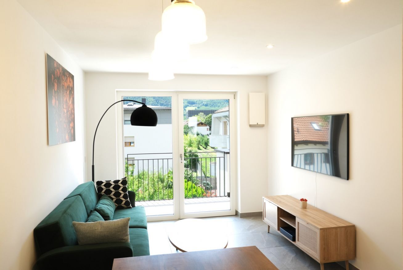 Ein modernes Wohnzimmer mit einem grünen Sofa, einem Holztisch, einer Stehlampe und einer Schiebetür, die auf einen Balkon mit Blick auf Grünflächen und Gebäude führt.