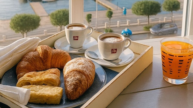 Un plateau de petit-déjeuner avec des croissants et des tasses de café, un verre de jus d’orange et une vue sur une marina, visible à travers une fenêtre.
