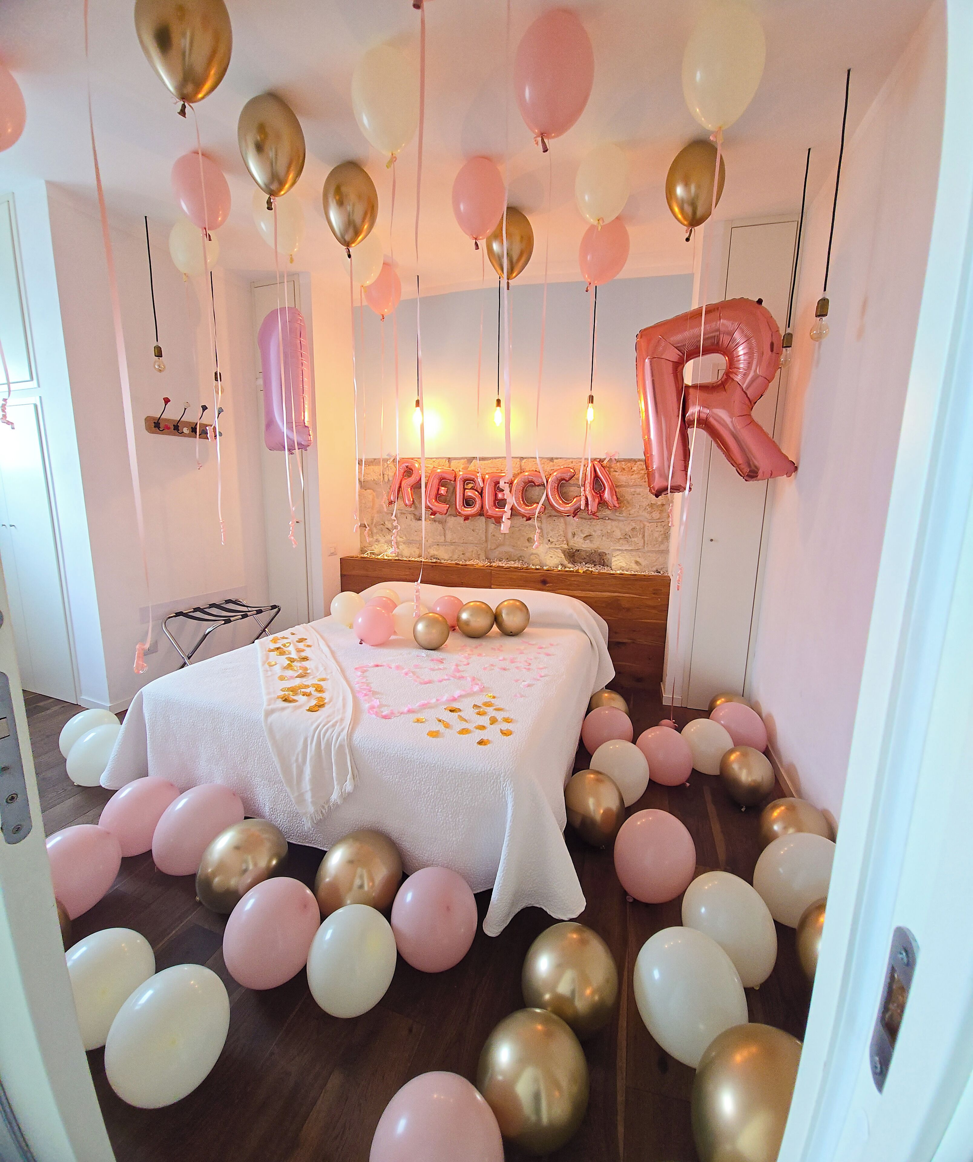 Une pièce avec un lit, des ballons et un mur sur lequel est écrit le nom « REBECCA » en lettres formées de ballons.