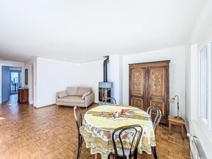 Ein Essbereich mit einem runden Tisch, der mit einer blumigen Tischdecke bedeckt ist und von Stühlen umgeben wird. Eine Schrankwand befindet sich an der Wand, und ein Holzschrank mit dekorativen Verzierungen ist zu sehen.