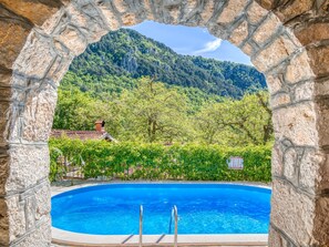 Una piscina con un arco de piedra, rodeada de abundante vegetación y con una montaña al fondo.