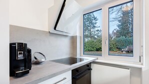 A modern kitchen with a built-in espresso machine, a window with a view of trees, and a white countertop. - Apartment 'Fewo Am Mühlenbach' with Shared Garden and Wi-Fi (Wehr)