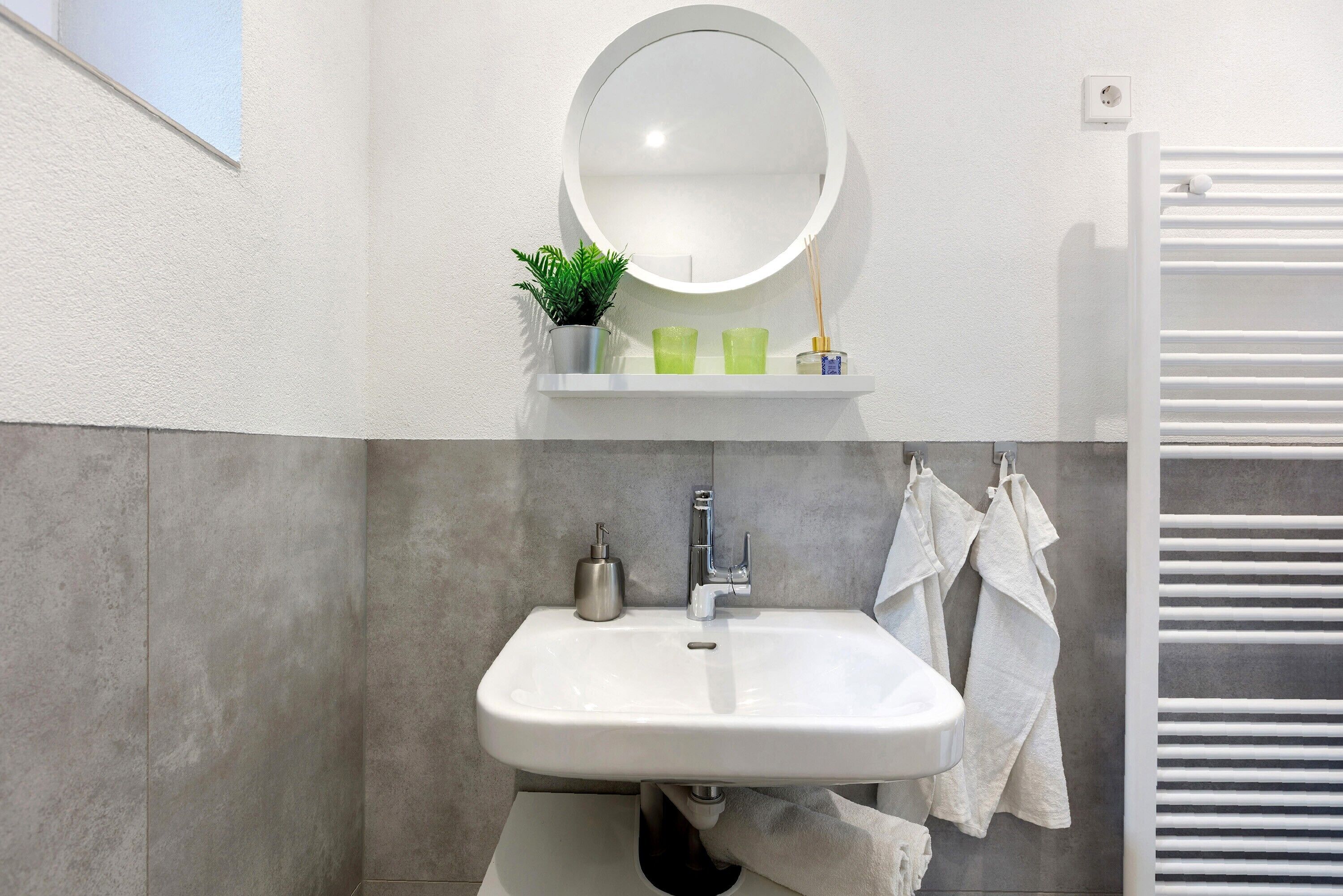 A bathroom with a white sink, a round mirror, a small shelf with a plant and two green glasses, and a towel rack with white towels.