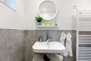 A bathroom with a white sink, a round mirror, a small shelf with a plant and two green glasses, and a towel rack with white towels. - Apartment 'Fewo Am Mühlenbach' with Shared Garden and Wi-Fi (Wehr)
