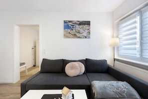 A modern living room with a grey sofa, a small table with a house model, a painting of a coastal scene, and a window with blinds.