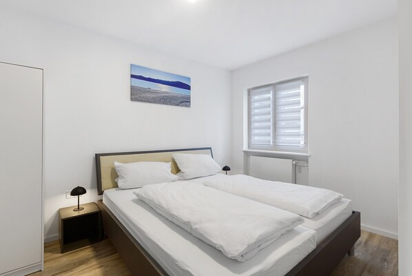 Une chambre à coucher avec un lit, une table de chevet, une lampe, une fenêtre et un tableau accroché au mur.