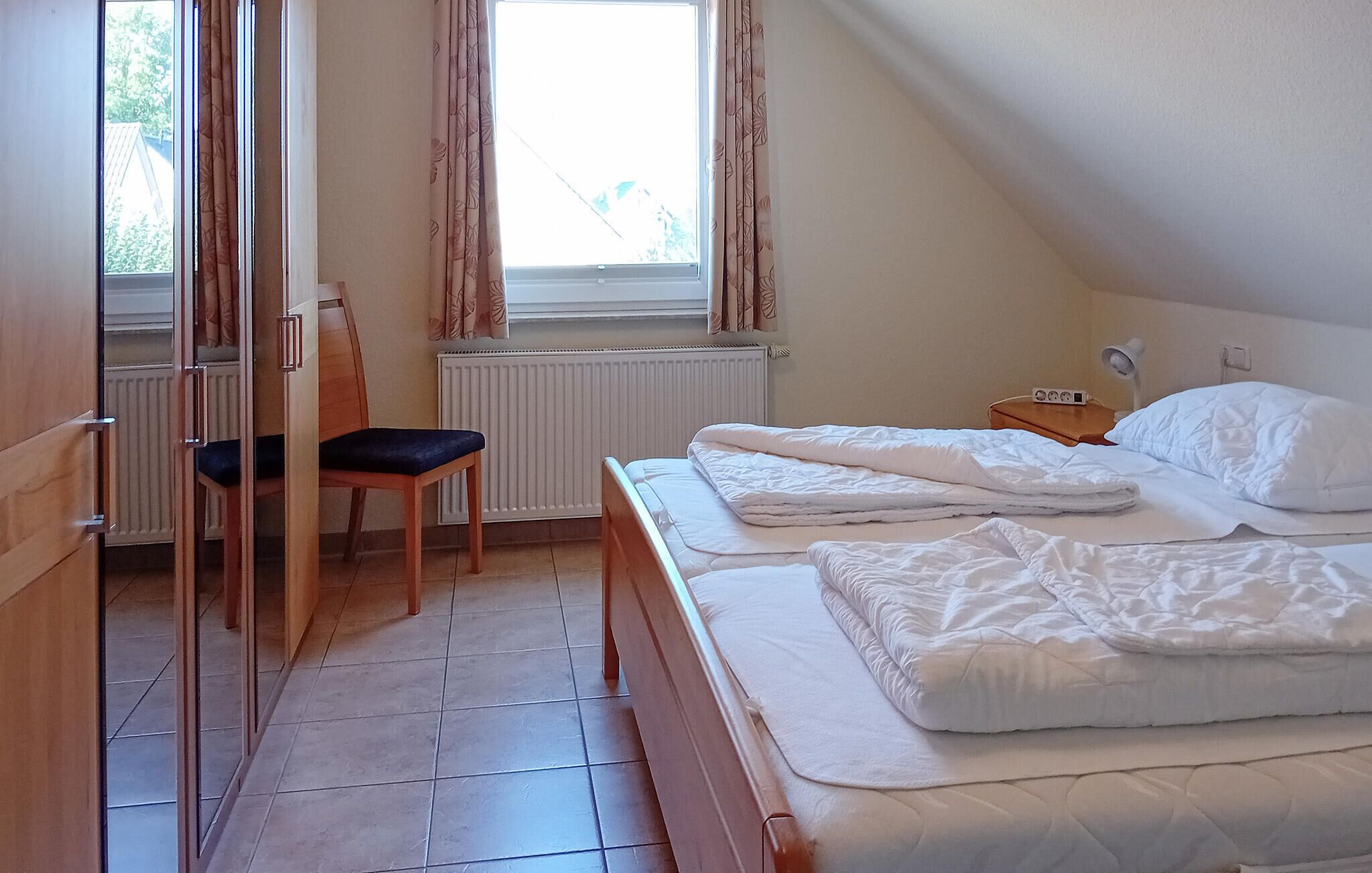 Un dormitorio con cama, una silla, una mesita de noche, una lámpara y una ventana con cortinas.