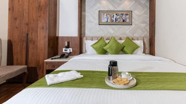 A hotel room with a bed, a nightstand, a wooden wardrobe, a framed picture on the wall, and a breakfast tray with a coffee pot and pastries. - Emerald Inn (Bhopal)