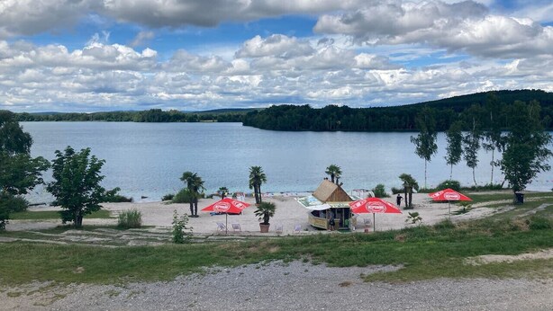 A lakeside beach with red umbrellas, a thatched-roof hut, and a clear view of the lake and surrounding hills. - Tiny house on Lake Murner with hot tub (Wackersdorf)