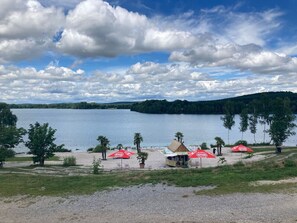 En strand ved søen med røde parasoller, en stråtækt hytte og et klart udsyn over søen og de omkringliggende bakker.