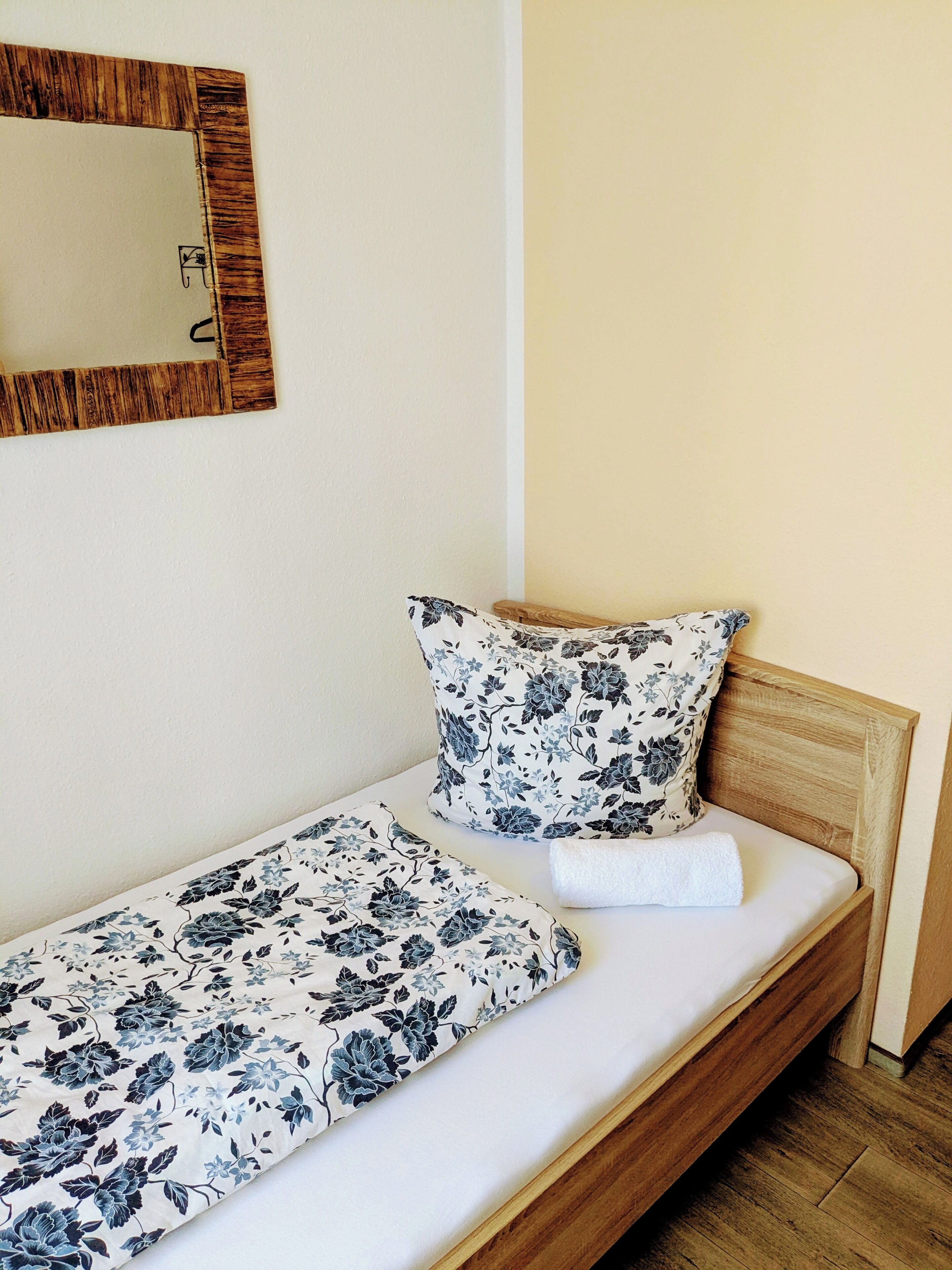 Une petite chambre avec un lit simple, une tête de lit en bois, une couvre-lit à motifs floraux et des coussins assortis. Un miroir à cadre en bois est accroché au mur, au-dessus du lit.