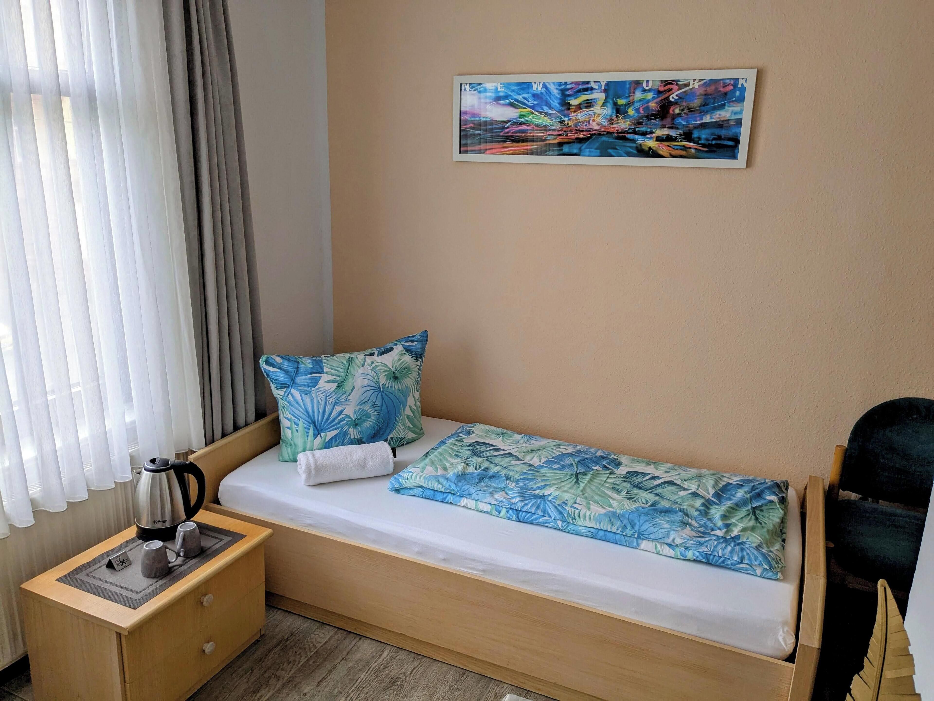 Una habitación pequeña con una cama individual, una mesita de noche de madera, una tetera y dos tazas. En la pared hay una almohada con motivos tropicales y un cuadro de un paisaje urbano.
