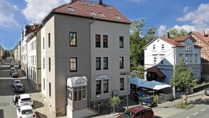 En toetages bygning med et skilt, der viser "Krombacher" i stueetagen, parkerede biler på gaden og en klar blå himmel.