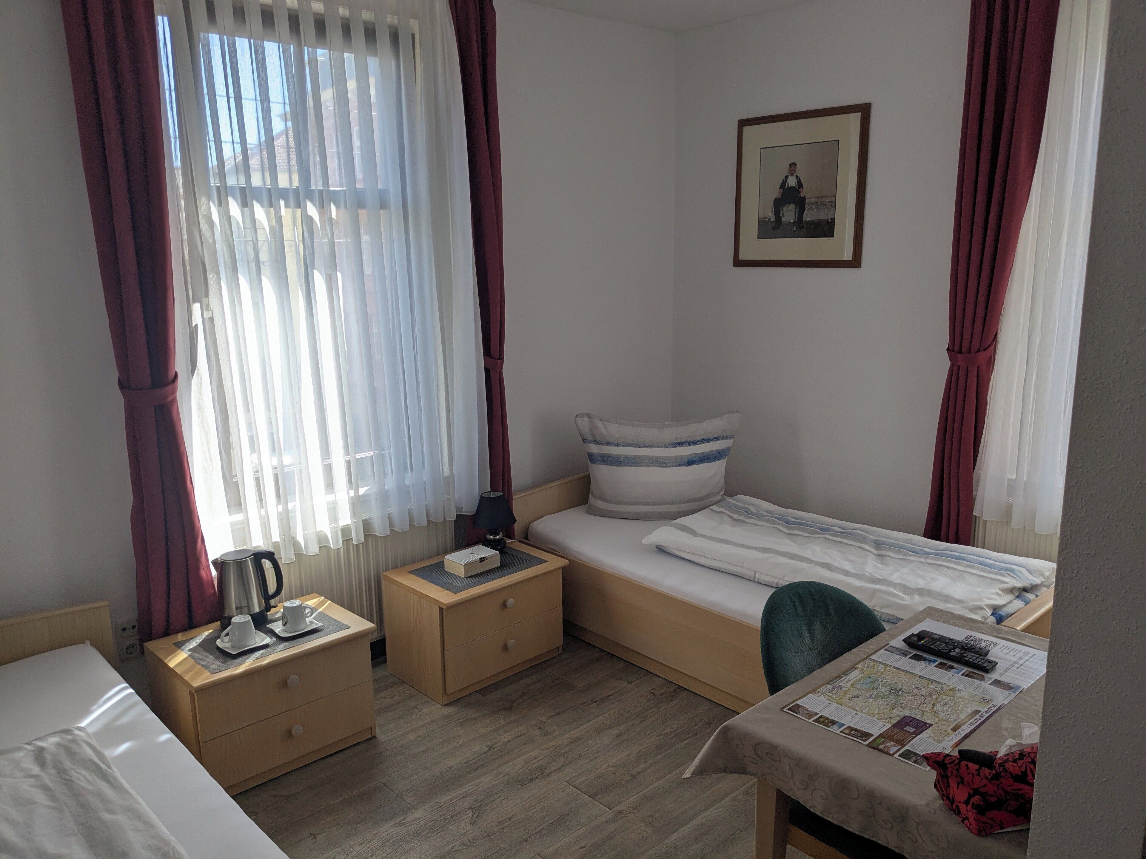 Un dormitorio pequeño y ordenado con una cama individual, una mesita de noche con una lámpara, una silla y una mesa con un mapa y mandos a distancia.