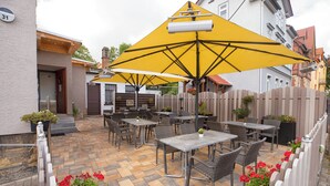En udendørs terrassemiljø med borde, stole, gule parasoller og krukker med planter.